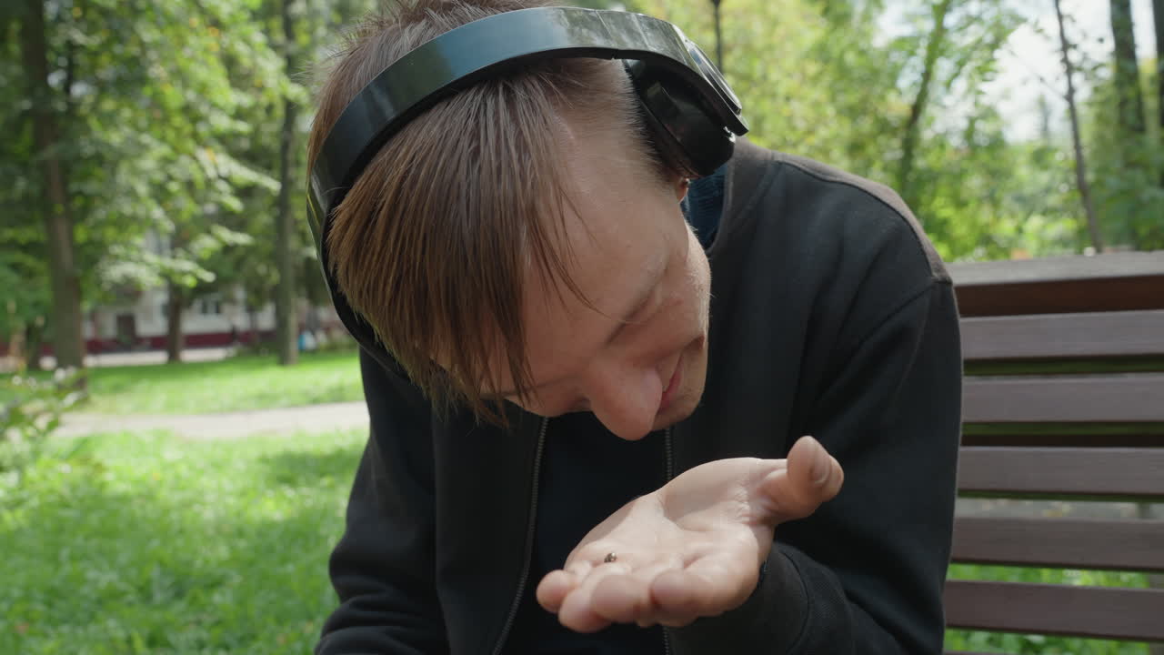 giovane uomo seduto su una panchina del parco che tiene un piccolo oggetto nel palmo della mano, indossa cuffie over-ear, luce solare soffusa tra gli alberi, ritratto spontaneo di curiosità, espressione gentile, luce naturale cinematografica, riconoscibile