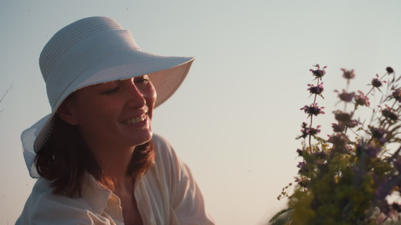 woman in white sunhat smiling brightly while admiring wildflower bouquet in soft evening light, warm tones enhancing joy and tenderness of serene outdoor moment surrounded by nature