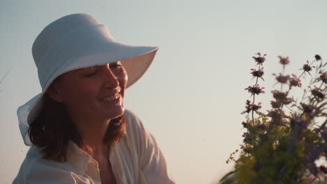 woman in white sunhat smiling brightly while admiring wildflower bouquet in soft evening light, warm tones enhancing joy and tenderness of serene outdoor moment surrounded by nature