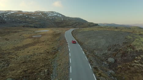 bright red car driving on a narrow, deserted road on a barren mountain in norway, drone shot with forward movement