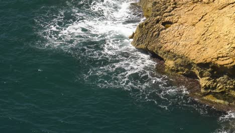 Rocky-headland-with-blue-ocean-waves-breaking-in-white-foam,-slow-motion