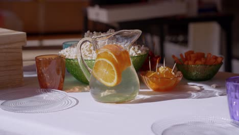 Pitcher-of-water-with-orange-on-table-for-celebration-with-fruit,-crudités-and-popcorn,-on-white-tablecloth