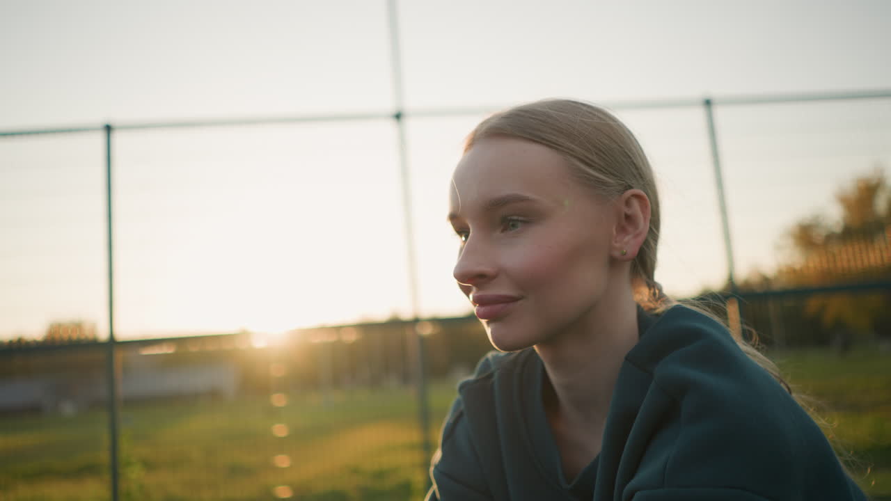 Young lady in green hoodie engaged in outdoor sport activity, sunlight creating a golden glow around her shoulders with open field and fence in the background during golden hour