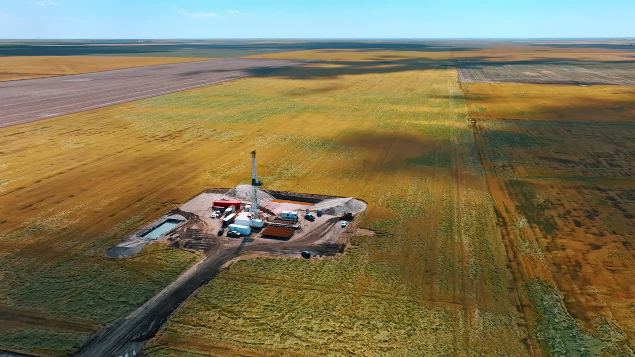 Flying closer to the site for oil production in the vast field. Several trucks surround the tower drilling the natural resources.