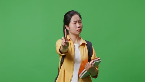 asian woman student with a backpack and some books shaking head and disapproving with no index finger sign while standing in the green screen background studio