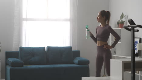 mujer deportiva está bebiendo agua durante el descanso en casa entrenamiento relajándose en la sala de estar en un día soleado actividad deportiva y estilo de vida saludable