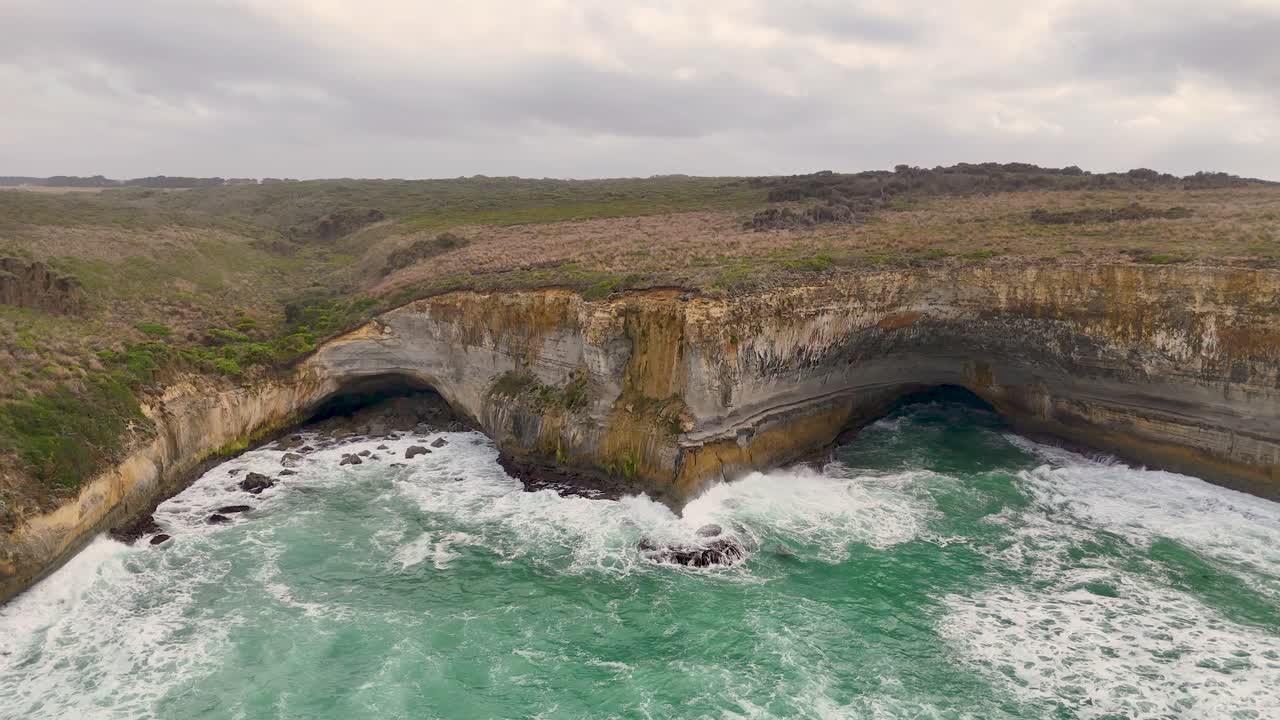 Drone footage captures the dramatic cliffs and turquoise waves of Port Campbell, Australia, under overcast skies, highlighting natural erosion and coastal beauty
