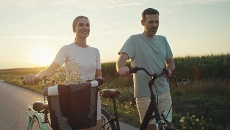 Fröhliches-Kaukasisches-Paar-Mittleren-Alters,-Das-Bei-Sonnenuntergang-Auf-Der-Dorfstraße-Mit-Dem-Fahrrad-Spaziert.