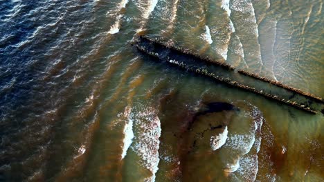 aerial-view-of-a-long-pier-jutting-out-into-a-turquoise-ocean-with-white-capped-waves-crashing-against-the-shore,-with-a-few-old-boats-bobbing-nearby