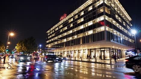 wet asphalt reflecting city lights, street lamps glowing, passing vehicles creating dynamic motion blur near contemporary glass facade building during nighttime urban landscape