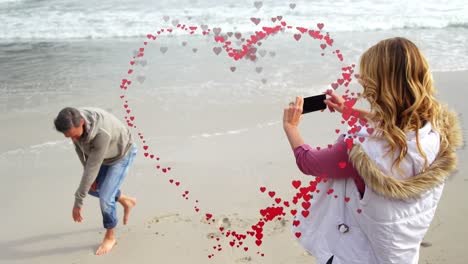 Animación-De-Pequeños-Corazones-Formando-Un-Corazón-Sobre-Una-Mujer-Caucásica-Filmando-A-Su-Novio-Saltando-En-La-Playa