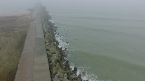 vista aérea del muelle de hormigón del puerto de liepaja, costa del mar báltico, día brumoso con niebla densa, sensación de mal humor, grandes olas de tormenta salpicando, amplios disparos de drones de ojo de pájaro avanzando