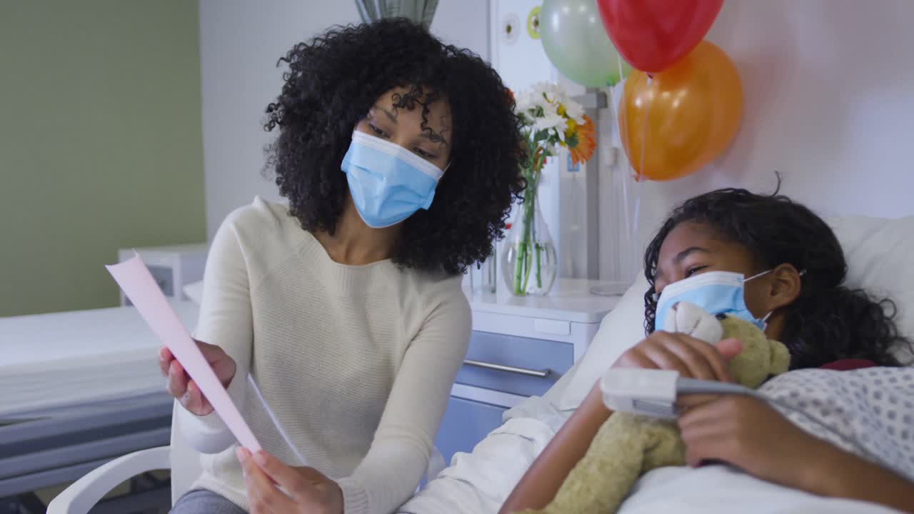 African american mother showing birthday card to her daughter lying on bed at hospital