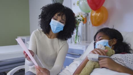 african american mother showing birthday card to her daughter lying on bed at hospital