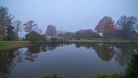 Lapso-De-Tiempo-Del-Amanecer-En-El-Lago-En-Un-Prado-Verde-Con-árboles-Y-Casas-De-Madera-En-Un-Día-Nublado-Y-Brumoso