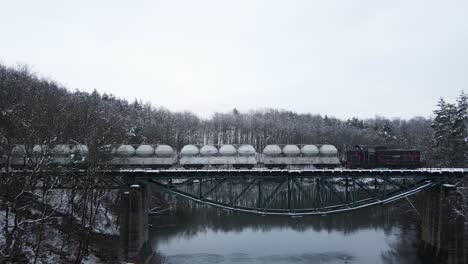 Tracking-Drohnenaufnahme-Eines-Frachtzugs,-Der-Eine-Brücke-über-Einen-See-Mitten-In-Einem-Verschneiten-Wald-In-Der-Kaschubei,-Polen,-überquert