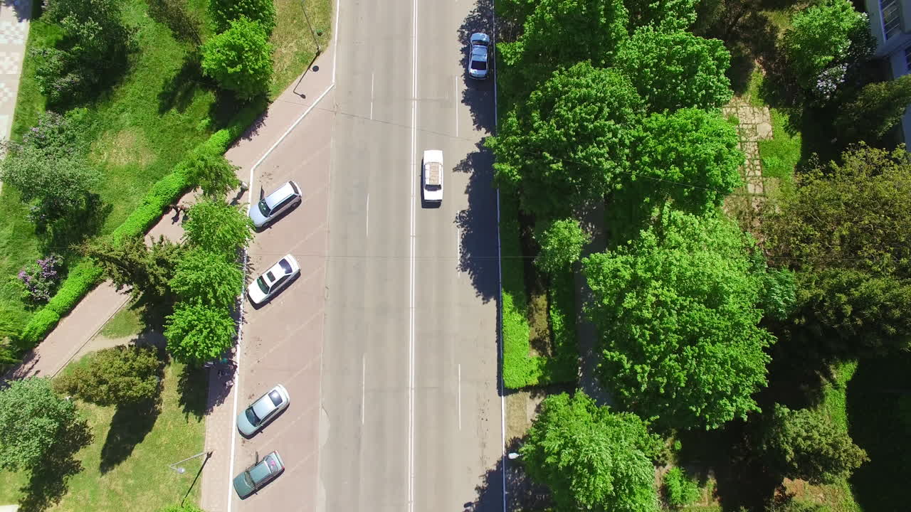 Following the white car going along the road surrounded by greenery. Residential area of a town from aerial perspective on sunny day. Top view.
