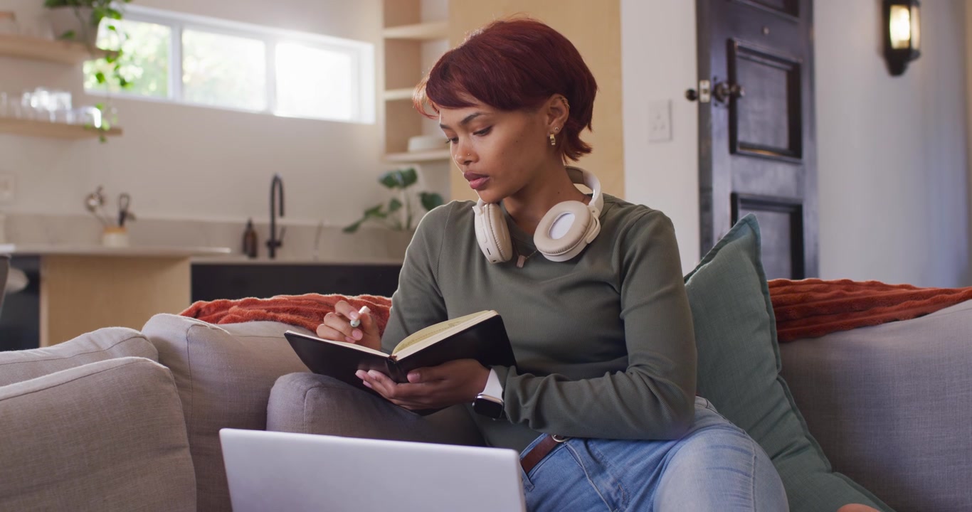 Adult African woman sitting on couch writing in notebook glancing at laptop white headset for study