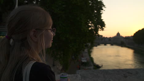 woman sips drink while looking over river in rome city at dusk, slow motion