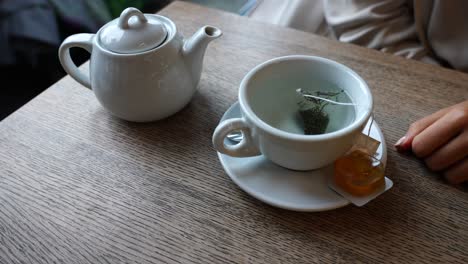 ceramic cup on wooden table with hot water and girl dip tea bag in it