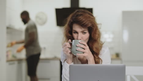 mujer blanca sonriente leyendo noticias felices en la computadora portátil en la cocina de la mañana.