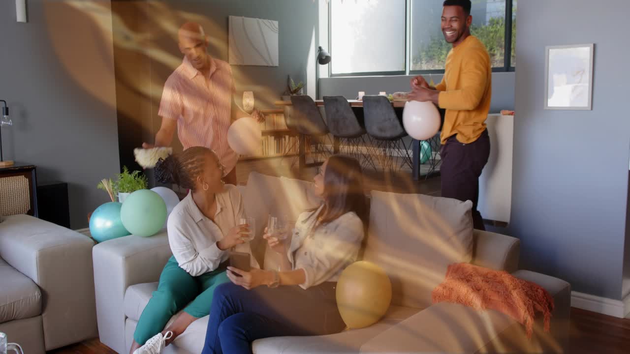 Seated pair raising phone for selfie as friends adding balloons, bowl, golden swirl capturing party