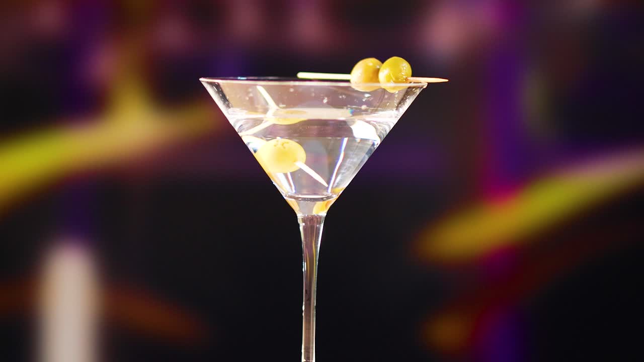 A hand places olives into a martini glass against a blurred, colorful bar background. Bright lighting highlights the cocktail