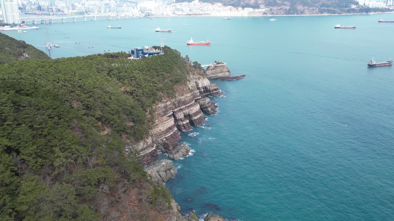 Drone aerial view in South Korea countryside flying over the clear blue sea of Busan next to a green mountain, ships and city in the background sunny day