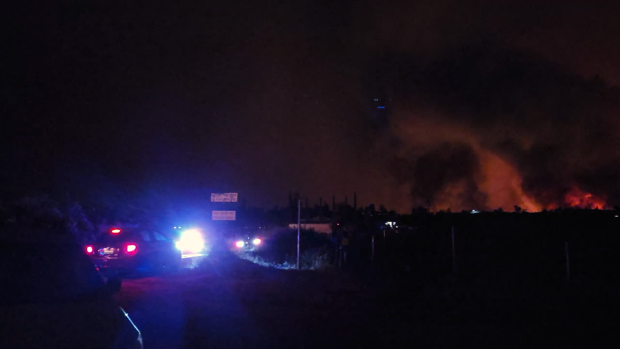 vehículos policiales patrullan cerca de incendios forestales por la noche, llamas rojas, columnas negras de humo se elevan