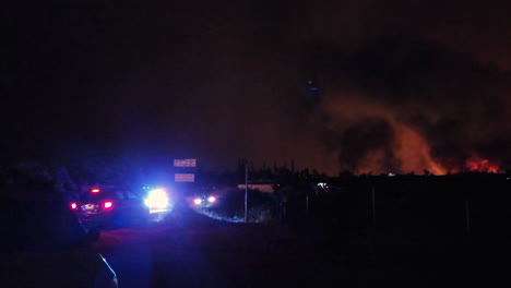 vehículos policiales patrullan cerca de incendios forestales por la noche, llamas rojas, columnas negras de humo se elevan