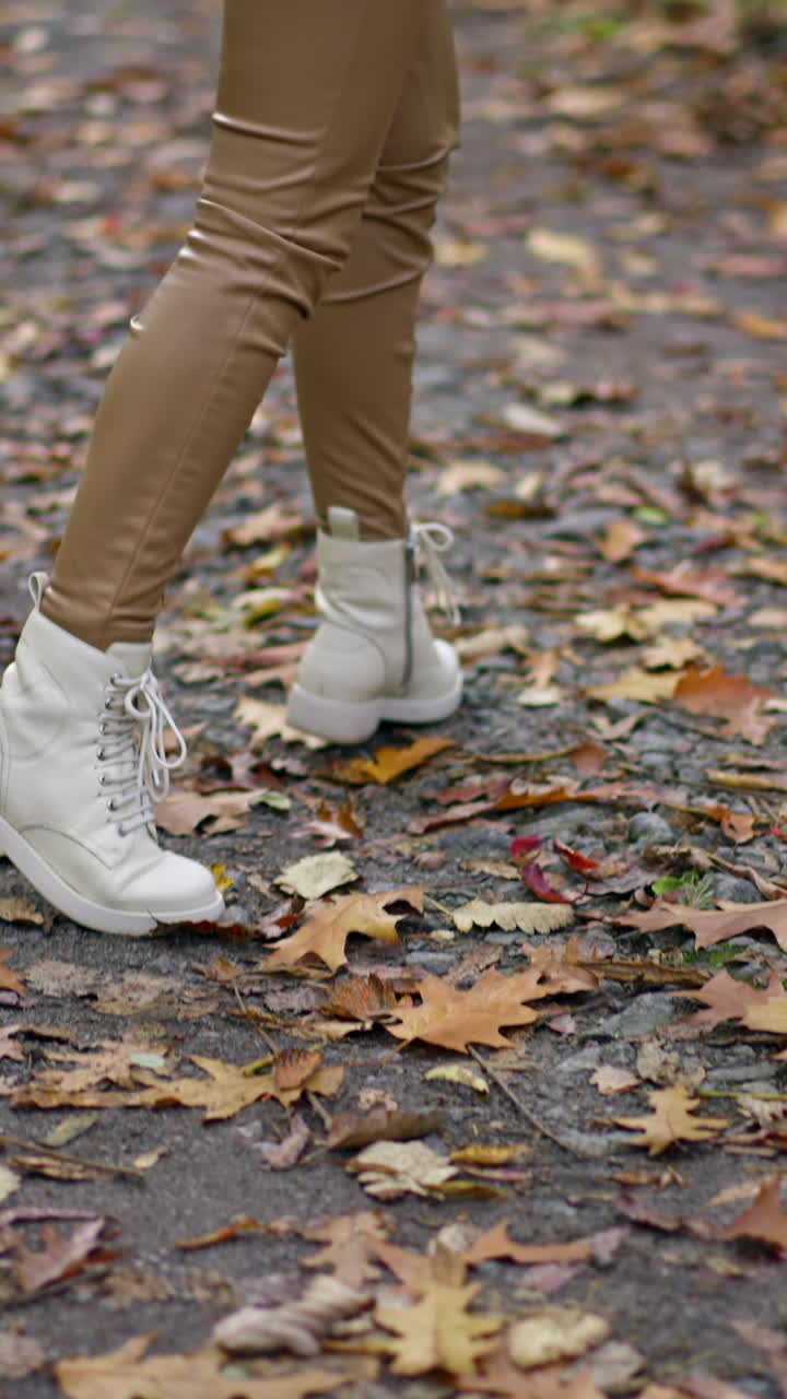 Unrecognized lady in beige pants and white jacket and boots walking in the park. Long-haired brunette turning around demonstrating her look.