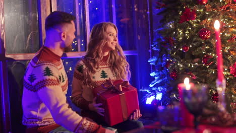 smiling couple sitting at the windowsill near a big christmas tree. woman is holding a present and man is kissing her.