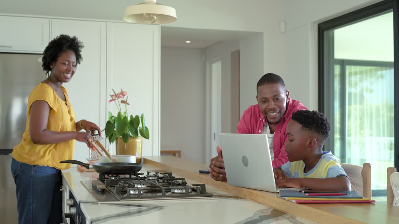 African American father guiding son typing on laptop in kitchen while mother stirring eggs at stove