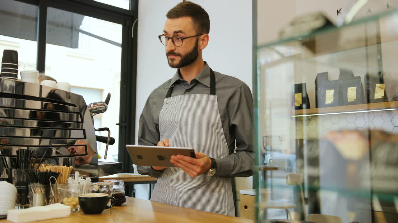 barista masculino caucásico con barba y gafas usando una tableta en una cafetería