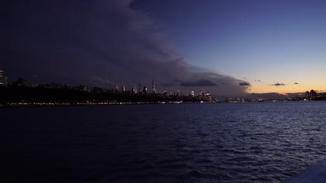 Horizonte-De-La-Puesta-Del-Sol-Del-Paisaje-Urbano-Moderno-De-La-Ciudad-De-Nueva-York-Iluminado-Al-Atardecer,-Barco-Acercándose-Al-Centro-De-La-Ciudad-Durante-La-Puesta-Del-Sol