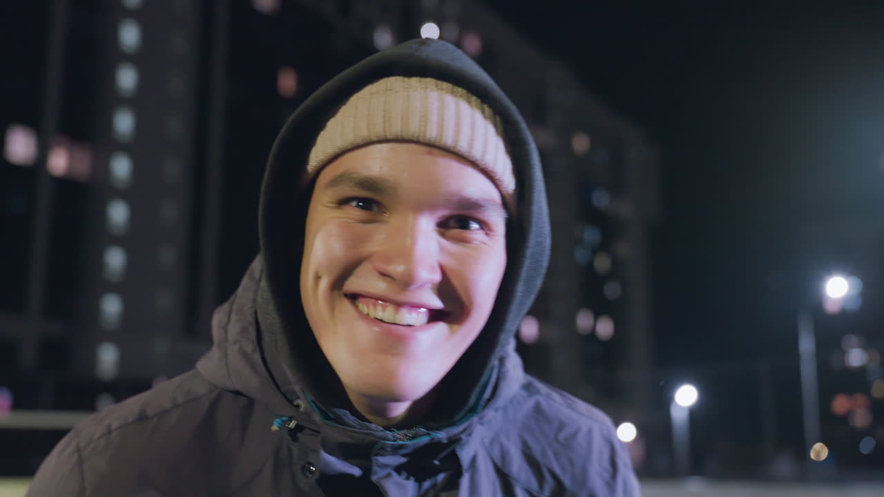 젊은 성인이 후디를 입고 밤에 도시 스포츠 경기장에서 축구기를 손에 들고 미소 짓고 있으며, 배경에는 조명된 주거 건물과 거리 조명이 있습니다.