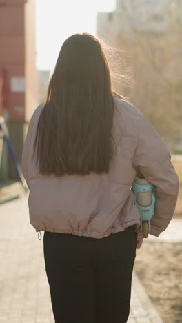 back shot of a girl wearing a peach jacket and black pants, holding a turquoise skateboard, walking on a sunlit pathway towards a building with sunlight streaming through the trees in the background