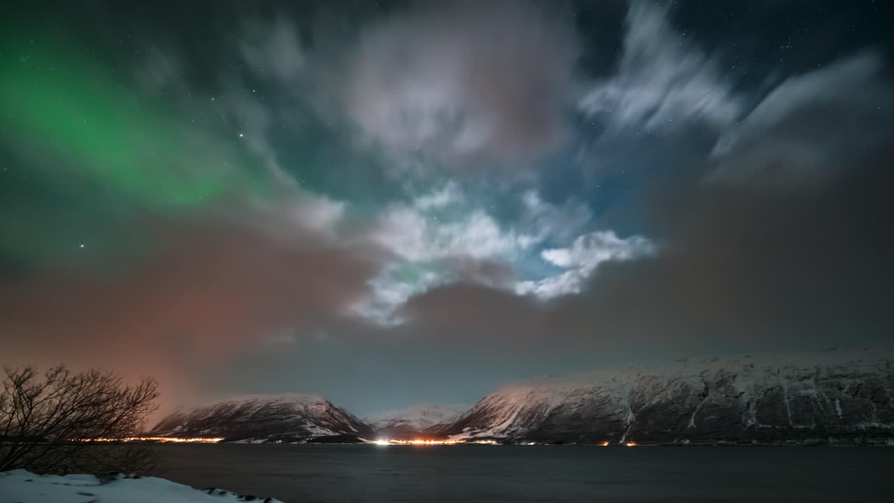 Snow-covered mountains and a fjord under a dynamic cloudy sky with northern lights, timelapse