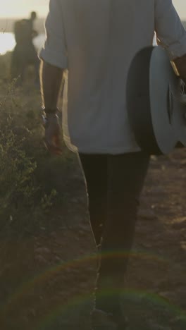 person walking on a dirt path carrying a guitar at sunset