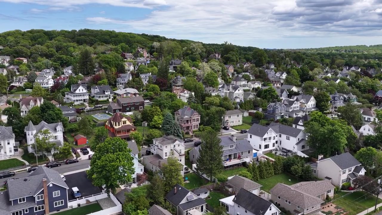 Monmouth County, New Jersey'deki Atlantic Highlands'ın havadan görünümü; ağaçlarla çevrili sokakları, müstakil evleri, yemyeşil bitki örtüsü ve parçalı bulutlu bir gökyüzünün altında dalgalı tepeleri olan bir banliyö mahallesini gösteriyor.