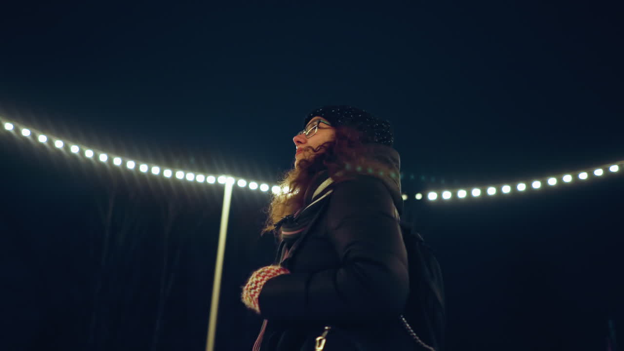 Woman in winter coat and knit hat standing under glowing string lights at night, gazing upward with peaceful expression, surrounded by cold dark sky and festive illumination creating dreamy atmosphere