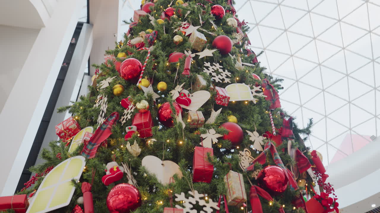 Close-up of tall, beautifully decorated Christmas tree with ornaments and lights in a modern mall, people walking in the background, festive shopping atmosphere with holiday cheer