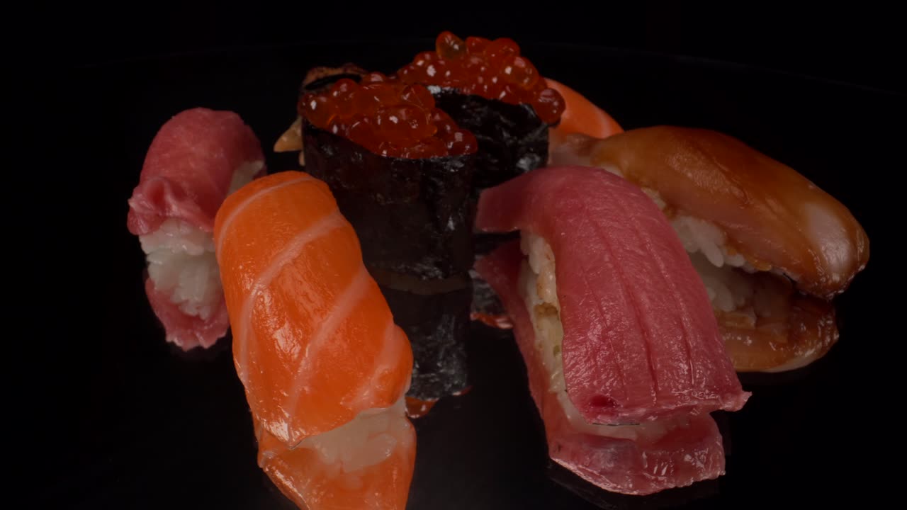 Makroansicht einer drehenden Nigiri-Sushi-Auswahl auf schwarzem, reflektierendem Glasuntergrund, mit Toro-Thunfisch, Lachs, Zitronenfisch, Lachsrogen und gegrilltem Aal, japanische Meeresfrüchte-Küche, 4K-Aufnahme.