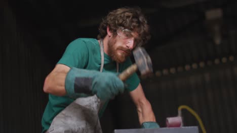 caucasian male blacksmith wearing safety glasses, hammering hot metal tool on anvil in workshop