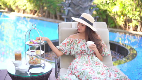 a cute woman seated on a poolside lounge chair enjoys a sip of her coffee as she reaches over to snag a sweet breakfast treat from the food caddy