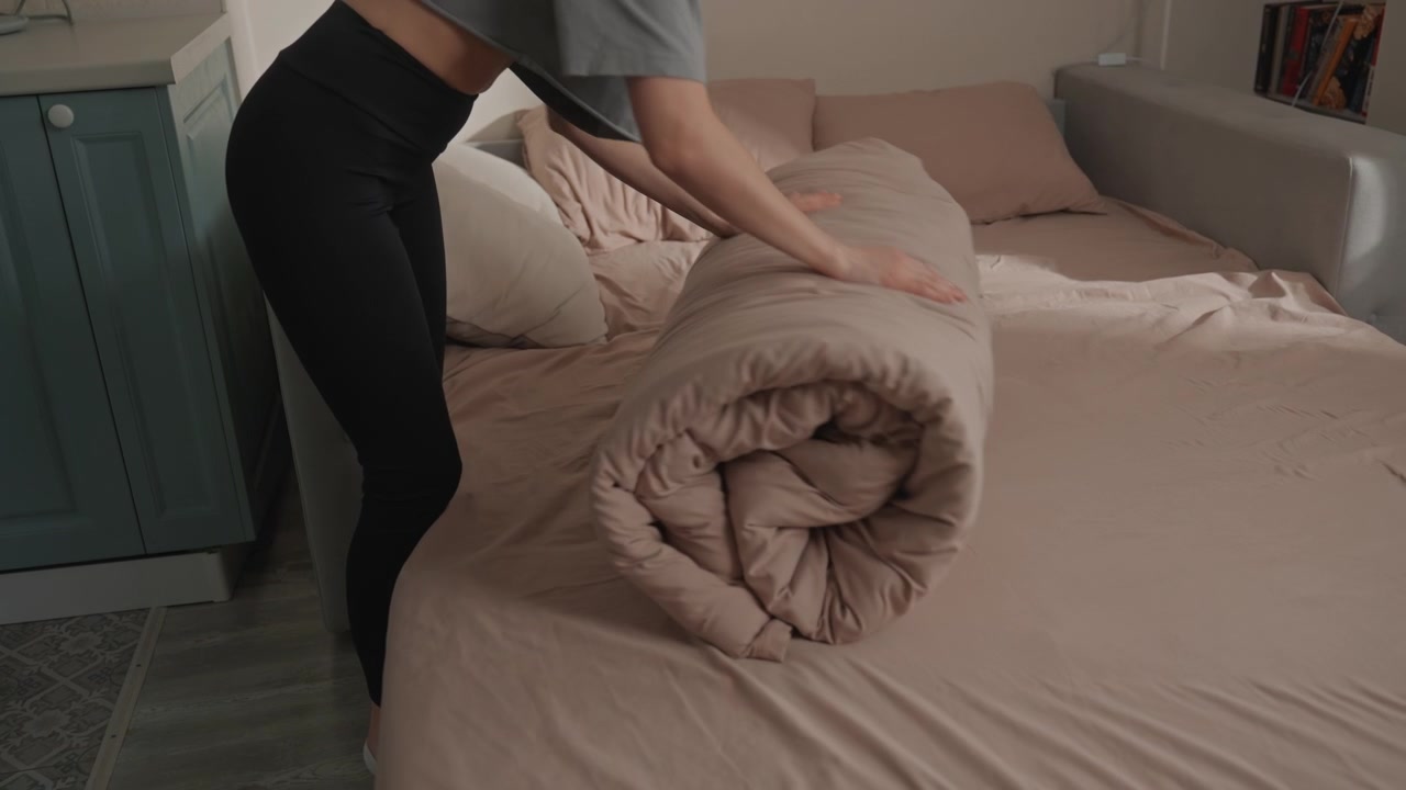 Wanita merapikan selimut di tempat tidur di kamar tidur minimalis, merapikan seprai dan menggulung selimut menjadi lipatan rapi, linen krem lembut, cahaya pagi yang hangat, koreografi domestik yang terfokus, lambat dan disengaja.