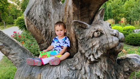 smiling caucasian kid sitting on the wooden sculpture of cat. happy toddler with a book outdoors.