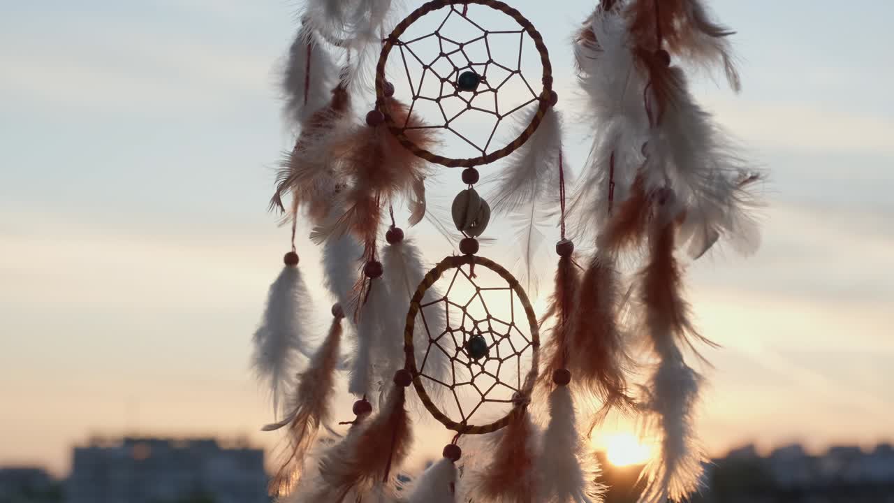 cazador de sueños con plumas naranjas gira, balanceándose con el viento ligero movimiento lento contra el fondo edificios de la ciudad de la noche, el cielo azul puesta de sol en verano. estilo boho, estilo indio. disco brillante del sol