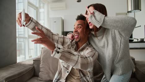 Ein-Glücklicher-Junger-Mann-Mit-Schwarzer-Hautfarbe-In-Einem-Karierten-Cremefarbenen-Hemd-Macht-Mit-Einem-Weißen-Smartphone-Ein-Selfie-Mit-Seiner-Brünetten-Freundin-Im-Pullover,-Während-Er-Auf-Einem-Modernen-Sofa-In-Einem-Studio-Apartment-Sitzt