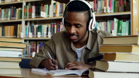 un guapo estudiante afroamericano con auriculares está escuchando música sentado en una mesa en una gran biblioteca espaciosa escribiendo conferencias rodeado de libros